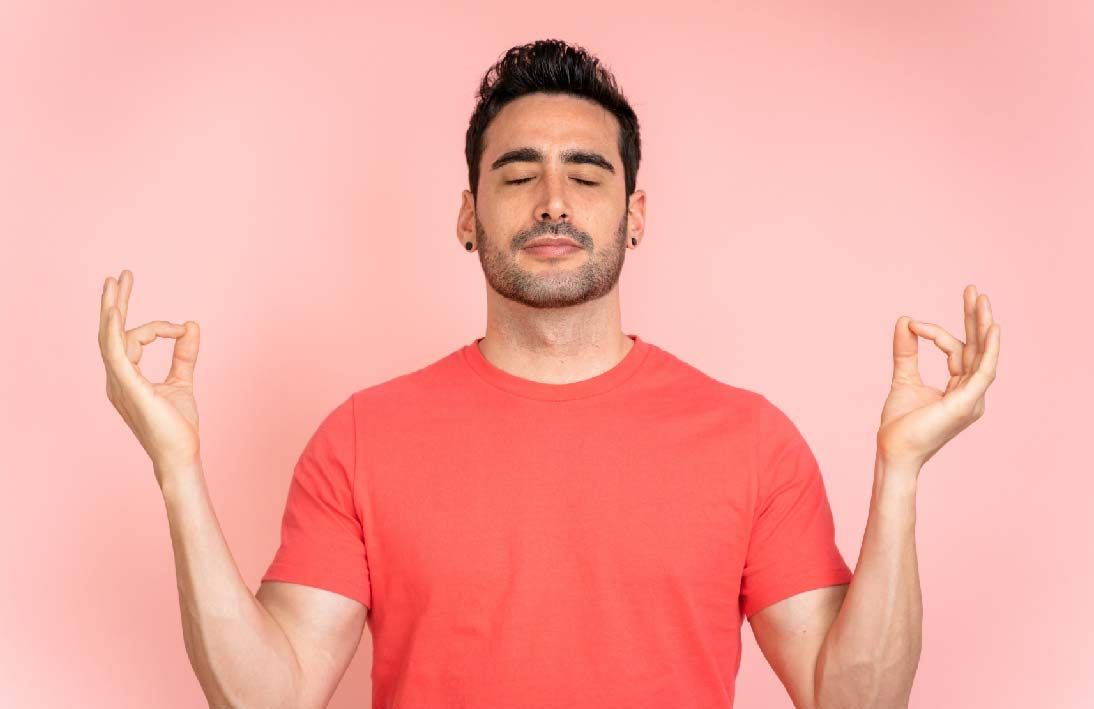 A man with dark brown hair wearing a pink shirt. Has his eyes closed with hands in a meditation pose.