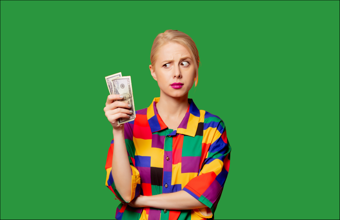 Blonde woman holding two notes of money, wearing a button up shirt with colourful squares or yellow, red, green, blue, and black. with one arm tucked under the arm holding the money while looking at the money confused.