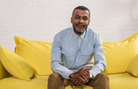 An older coloured man sitting on a yellow couch holding his hands with a slight smile on his face. Looking like he is waiting for a conversation with you.