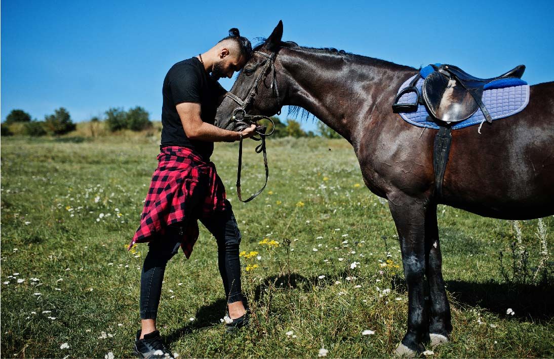 An Arab man wearing black with a red shirt tied around his waste, standing in a field holding a horses head while they both stand head to head together looking at peace.