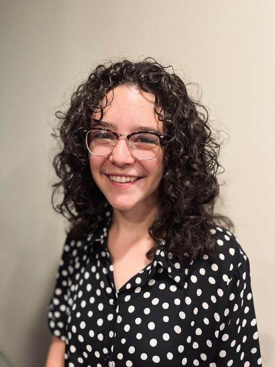 Hannah G smiling and welcoming standing in front of a light colored background.