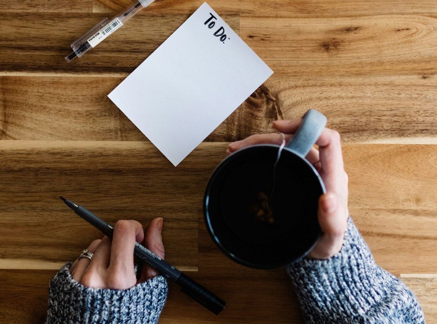 A person holding a cup of tea while writing a to‑do list at a wooden table.
