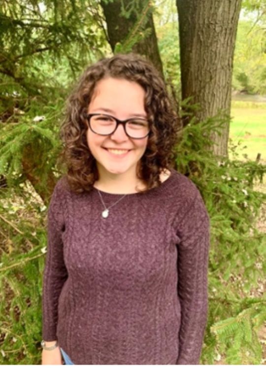 Hannah G smiling and welcoming standing in front of a light colored background.