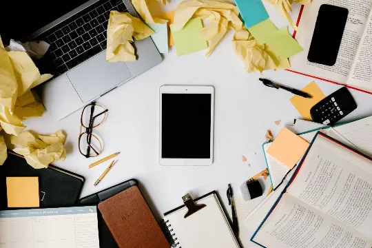 A busy desk with crumpled post it notes, a computer, a tablet, multiple notebooks all open to different pages.