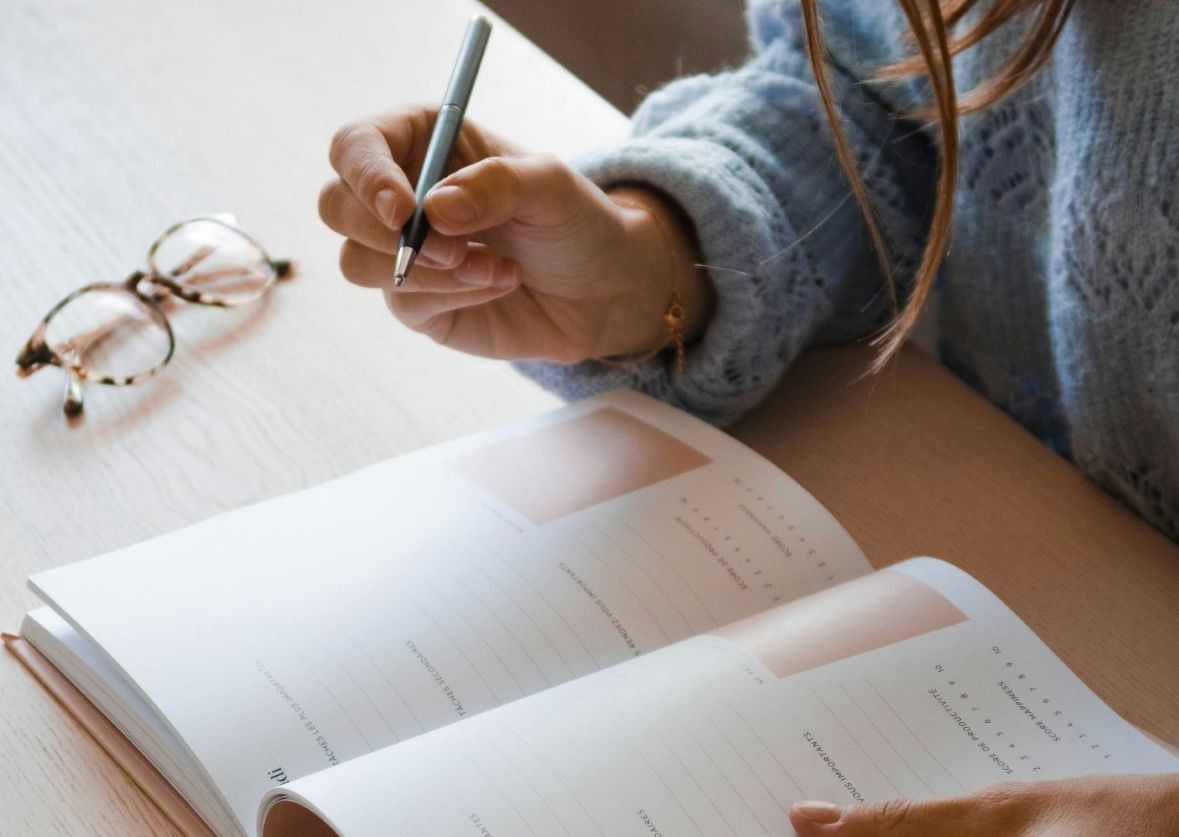 Hand gently holding a pen poised to write in a clean, neat planner with glasses neatly folded on the side